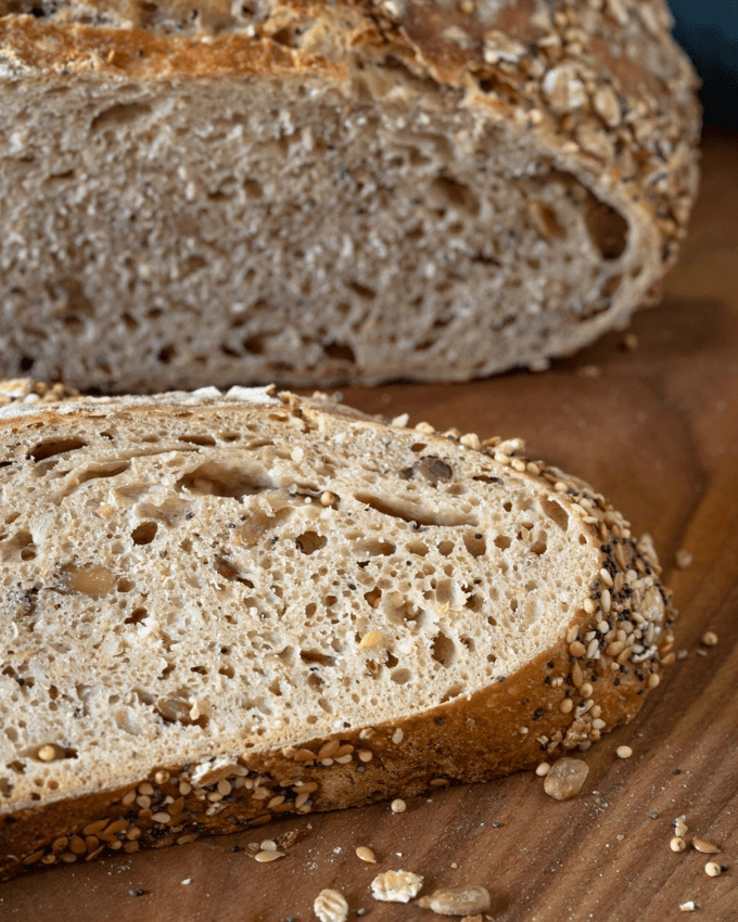 Close up angled photo of a slice of wheat bread with seeds and oats baked into it in front of the loaf.