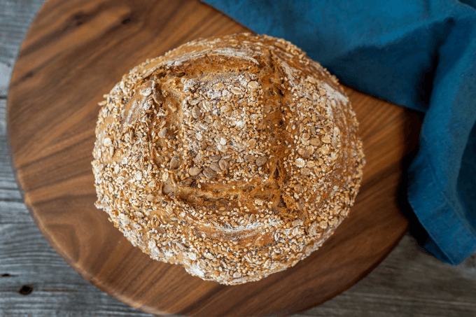 Top down photo of a loaf of Sourdough Multigrain bread with a multi-grain crust. It sits on a wooden cutting board next to a blue napkin.