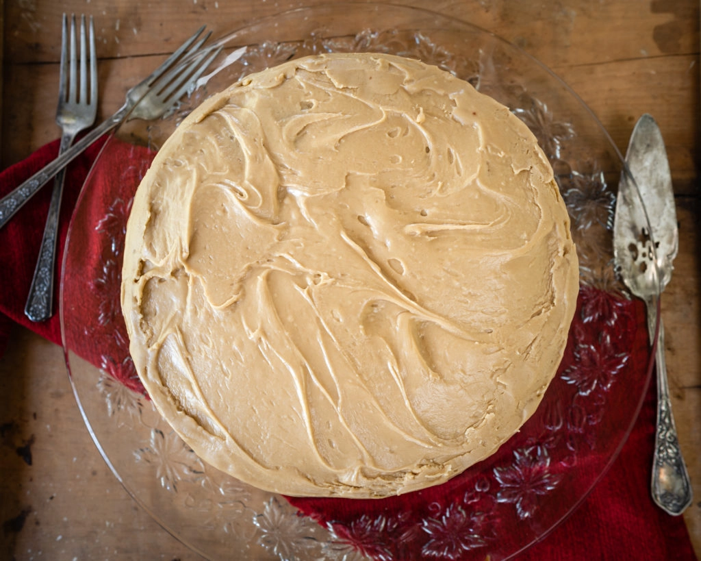 Top down view of a round layered cake on a glass platter over a red napkin on a wooden board. Antique cake servers and forks sit beside the cake.