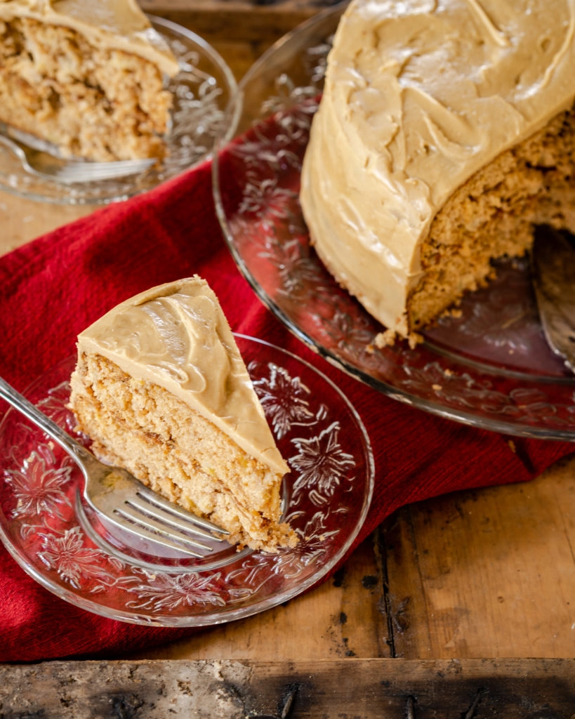 Top-down photo of a slice of apple cake next to the cut round cake on a red napkin sitting on a wooden board. A second slice sits in the background.