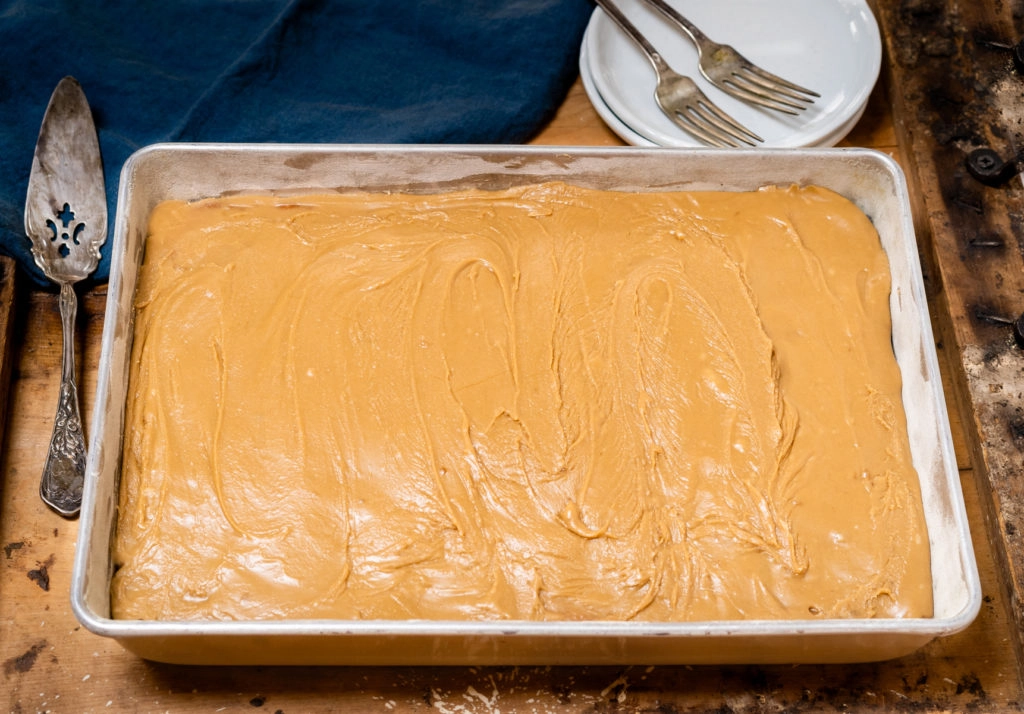 Top angled view of a 9x13 cake frosted with caramel icing sitting on a wooden board. Two serving plates with forks and a blue napkin sits behind it.