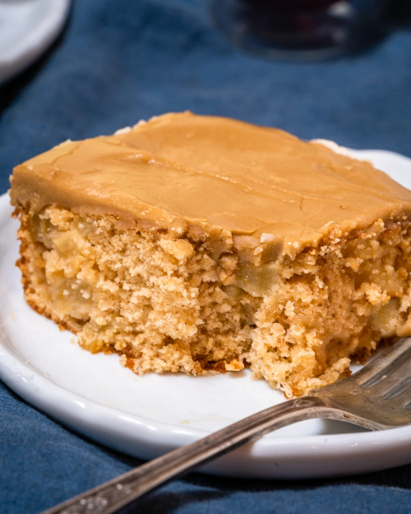 A slice of cake with a bite taken out of it showing chunks of apples inside and frosted with a caramel icing. It sits on a white plate next to a fork over a blue napkin.