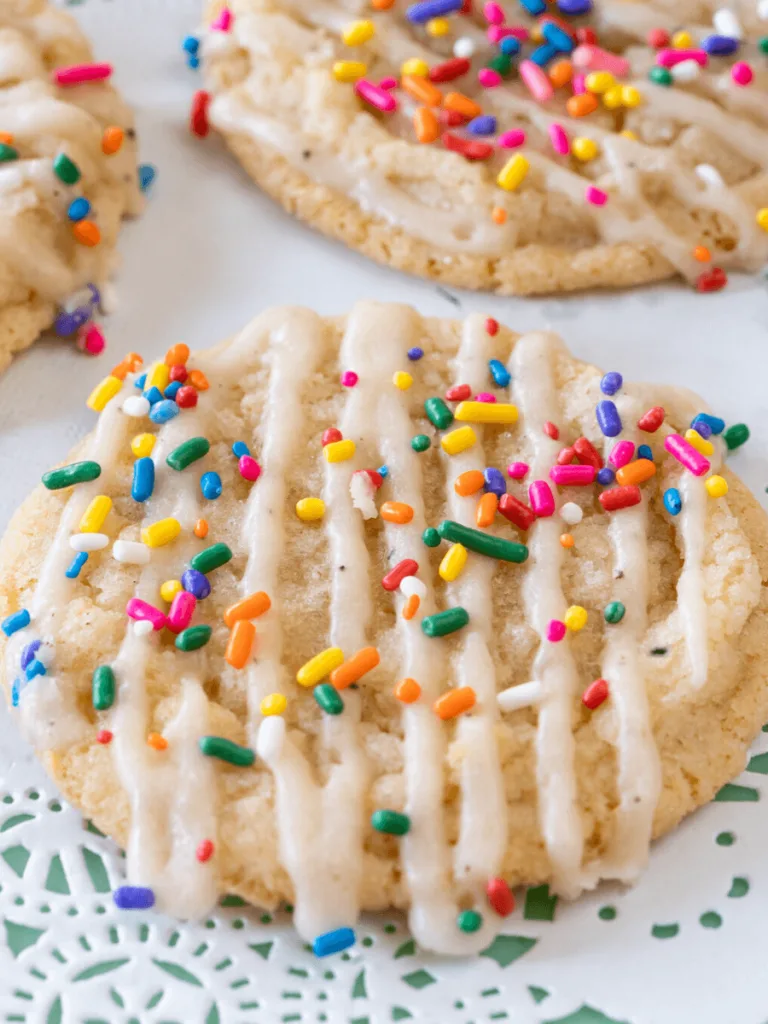 Close angle view of a sugar cookie with glaze piped in stripes across the top and colorful sprinkles on a paper lace tray.