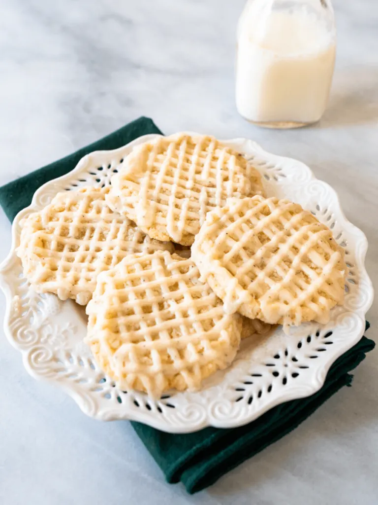 4 sugar cookies that have piped glaze in a hatch pattern on a white plate in front of a bottle of milk sitting on a marble background.