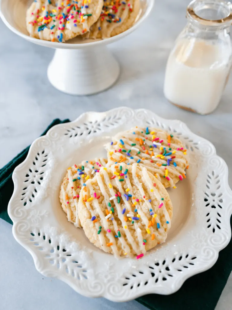 Top shot of 3 glazed and sprinkled cookies on a white plate in front of a bottle of milk and a pedestal dish with more cookies.