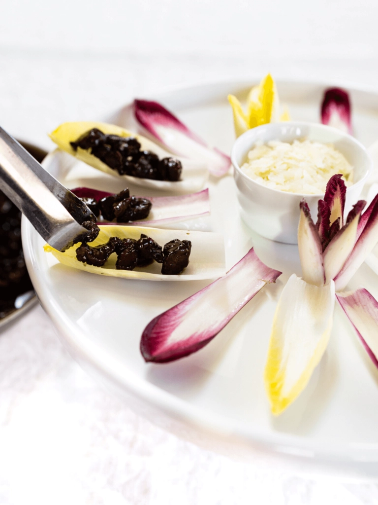 Tongs placing bits of lamb on endive lettuce leaves on a white serving platte with a cup of cheese in the center of the platter