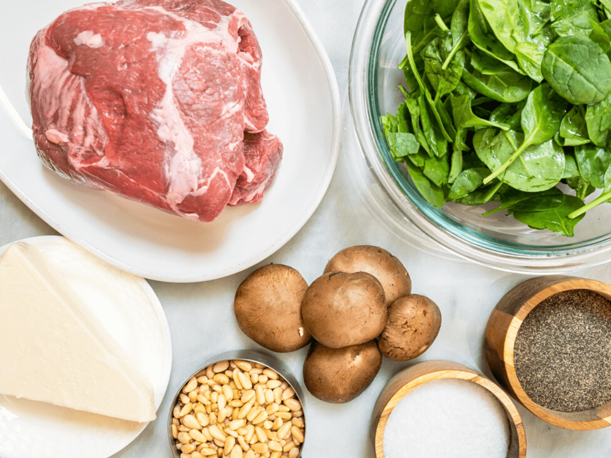 Top down view of the ingredients used to make lamb roulade. Leg of lamb, spinach, mushrooms, cheese, and pine nuts.