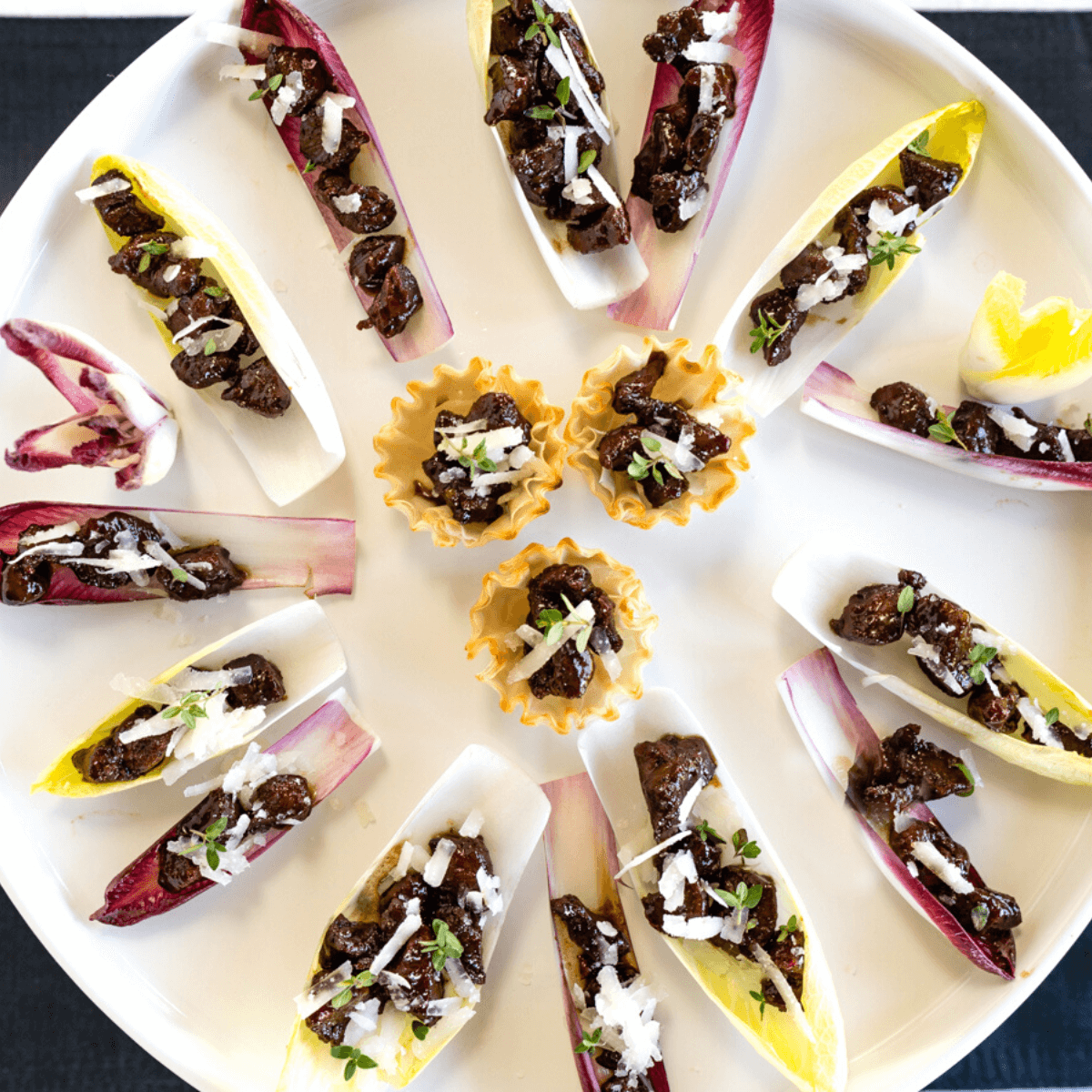 Top view of a round white platter filled with endive lettuce leaves and phyllo cups filled with lamb bits, pecorino cheese and fresh thyme