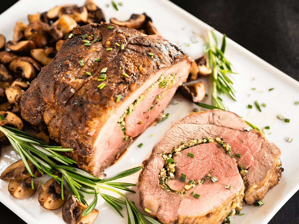 Top down view of a roasted leg of lamb that's been butterflied and rolled. It's stuffed with pinenuts, mushrooms, and spinach. Theres chopped mushrooms surrounding the roast on a white platter.