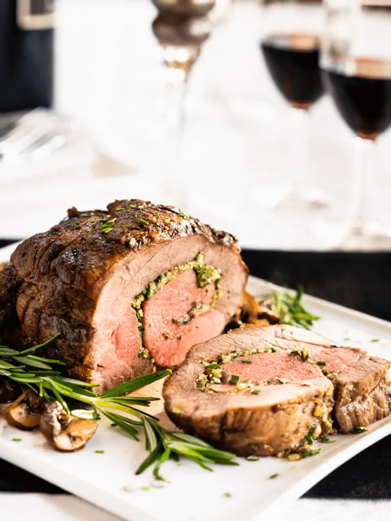 Straight on view of a roasted lamb roulade stuffed with a filling of pine nuts, cheese, mushrooms, and spinach on a white plate surrounded by mushrooms and rosemary sprigs.