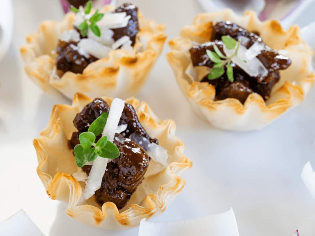 Top down view of 3 phyllo cups stuffied with lamb bits drizzled with fig balsamic glaze and sprinkled with pecorino and fresh thyme on a white plate.