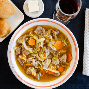 Top down photo of a bowl of vegetable lamb soup in a white bowl sitting on a black placemat. A glass of wine and plate of bread and butter sit behind it.