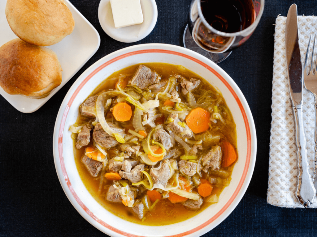 Top down photo of a bowl of vegetable lamb soup in a white bowl sitting on a black placemat. A glass of wine and plate of bread and butter sit behind it.