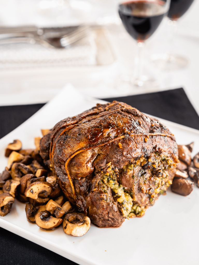 Roasted lamb roulade straight from the oven before it\'s sliced. It sits next to a bed of roasted mushrooms on a white plate over a black placemat.