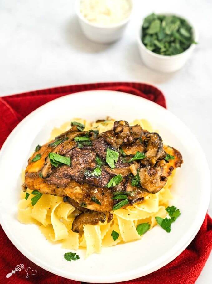 Angled view of a dish of browned chicken breasts on a bed of egg noodle pasta covered with a creamy mushroom marsala sauce and garnished with parsley in a white bowl sitting on a red napkin. with small dishes of parsley and cheese in the background.