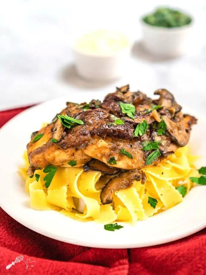 Side view of a dish of browned chicken breasts on a bed of egg noodle pasta covered with a creamy mushroom marsala sauce and garnished with parsley in a white bowl sitting on a red napkin. with small dishes of parsley and cheese in the background.