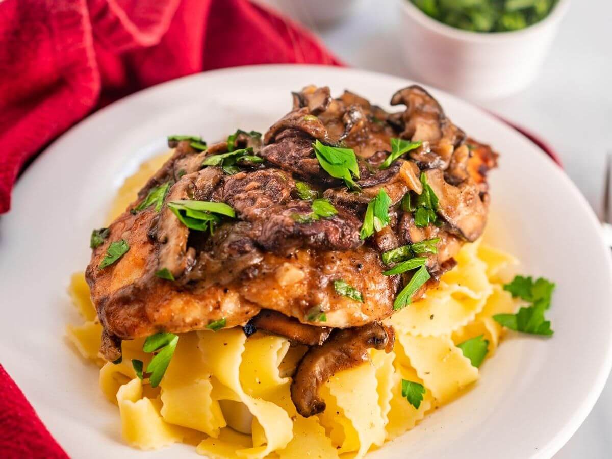 Side view of a dish of browned chicken breasts on a bed of egg noodle pasta covered with a creamy mushroom marsala sauce and garnished with parsley in a white bowl sitting on a red napkin. with small dishes of parsley and cheese in the background.