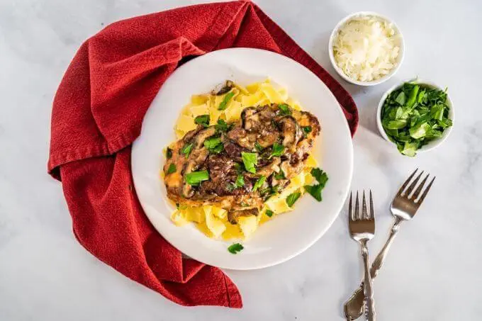 Top down view of a dish of browned chicken breasts on a bed of egg noodle pasta covered with a creamy mushroom marsala sauce and garnished with parsley in a white bowl sitting on a red napkin. with small dishes of parsley and cheese in the background.