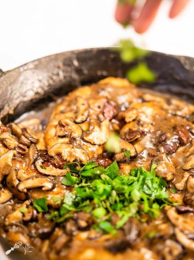 A photo of parsley being sprinkled over the chicken covered with a marsala sauce in a cast-iron skillet