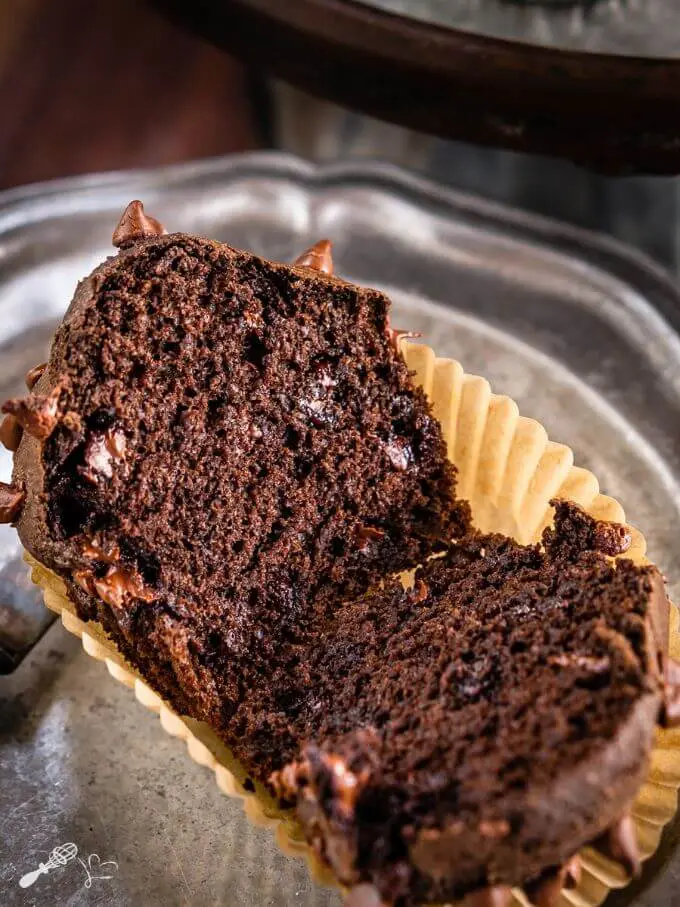 Double Chocolate Muffin sliced in half showing a soft inner crumb and melted chocolate chips. The muffin sits in a tan muffin paper on a silver tray.