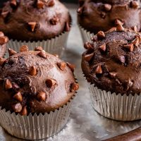 Close up side view of two in focus chocolate muffins dotted with chocolate chips sitting on a silver tray with 3 out of focus muffins sitting in the back of the tray.
