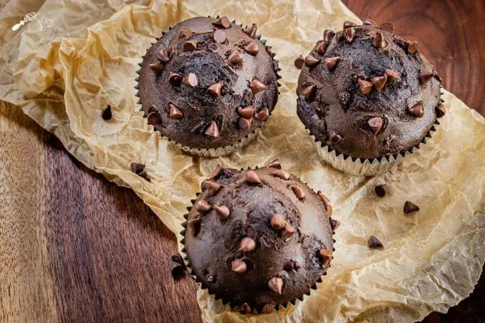 Top looking down at three chocolate muffins sitting on wrinkle parchment paper over a wooden cutting board.