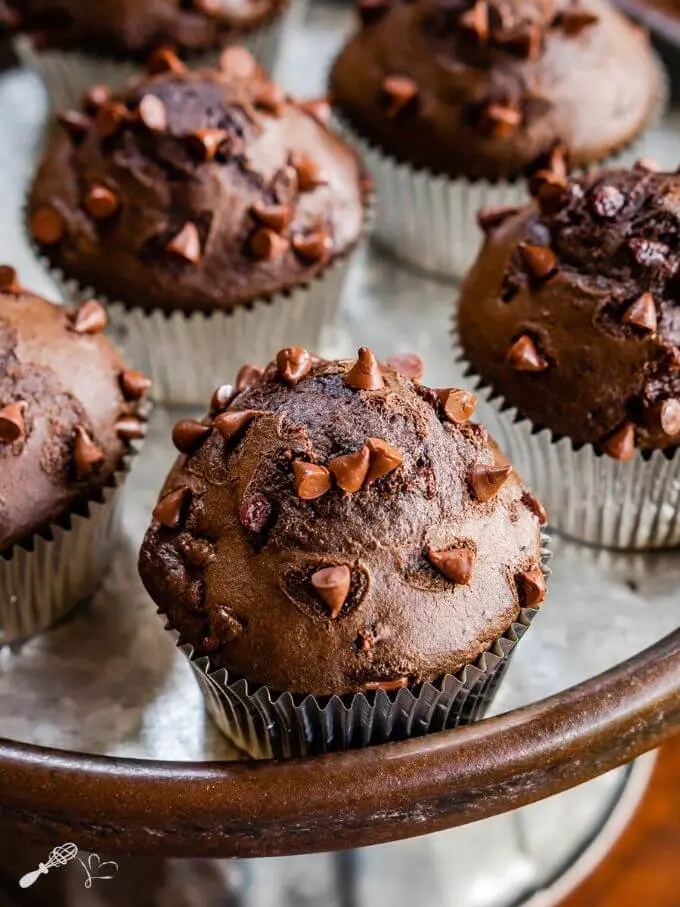 Silver pedestal tray topped with a double chocolate muffin with chocolate chips on top in front of 4 out of focus muffins.