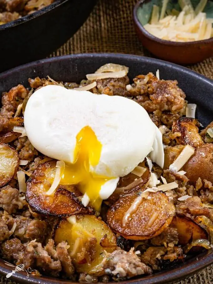 Top down photo of sausage and fried potatoes sprinkled with parmesan cheese with a soft cook egg pouring over it.