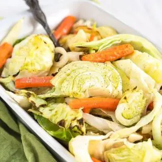 White baking dish filled with roasted green cabbage, orange carrots, apples and onions with a silver serving spoon in the pan sitting on a green napkin.