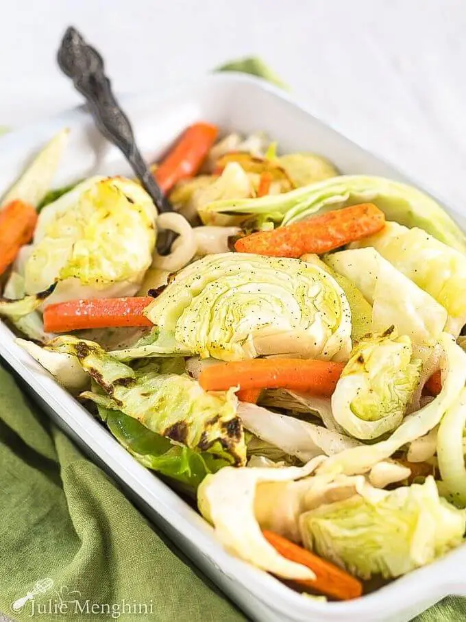 White baking dish filled with roasted green cabbage, orange carrots, apples and onions with a silver serving spoon in the pan sitting on a green napkin.