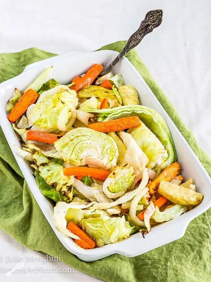 White baking dish filled with roasted green cabbage, orange carrots, apples and onions with a silver serving spoon in the pan sitting on a green napkin.