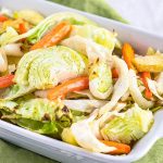 White baking dish filled with roasted green cabbage, orange carrots, apples and onions with a silver serving spoon in the pan sitting on a green napkin.