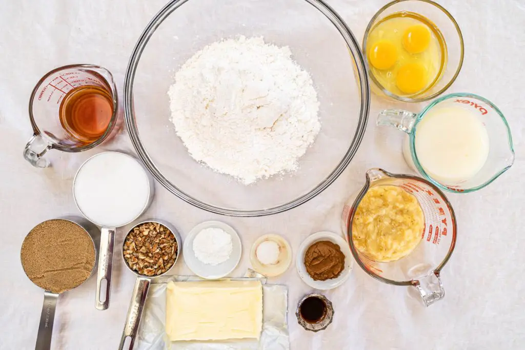 Bowls of the ingredients necessary to make a bananas foster cake including flour, sugar, brown sugar, rum, buttermilk, eggs, vanilla, baking powder, salt, cinnamon, butter, and pecans.