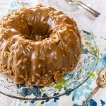 Top angle of a bananas foster bundt cake frosted with a caramel pecan praline glaze on a glass plate over a blue floral napkin. Plates, a cake server, and forks sit in the background.