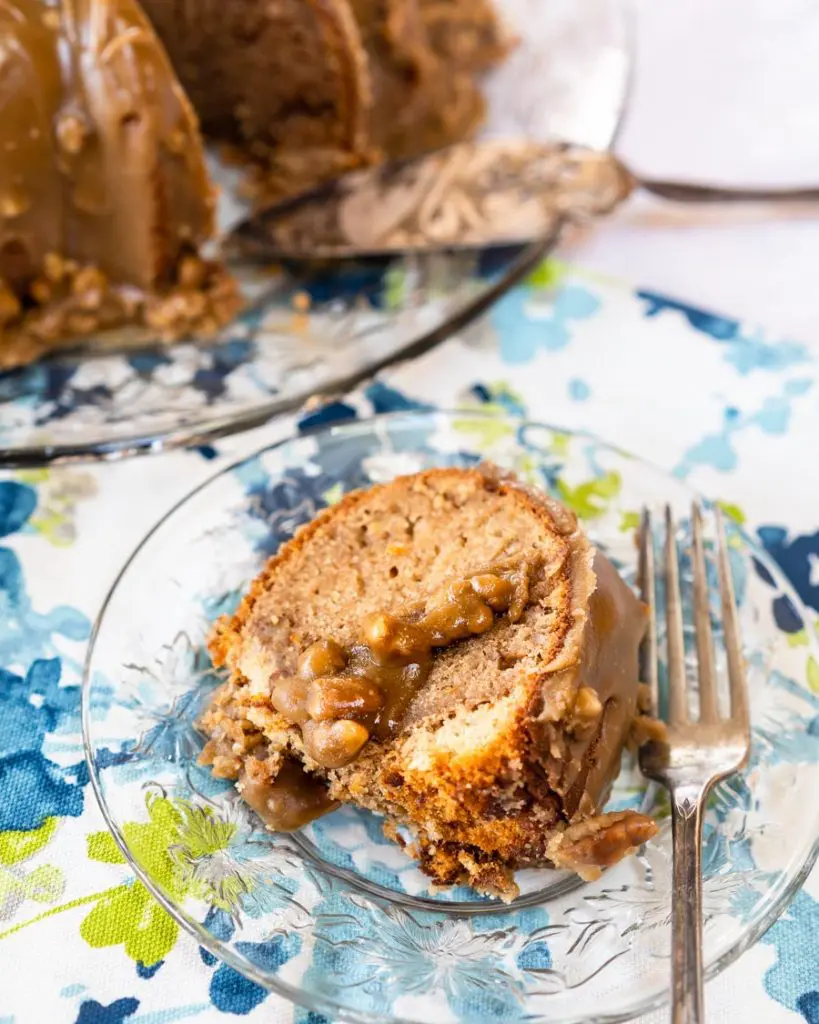 A slice of a bananas foster bundt cake drizzled with pecan praline glaze sitting on a glass plate over a blue floral napkin. A fork sits on the plate next to the cake. The cake sits in the background.