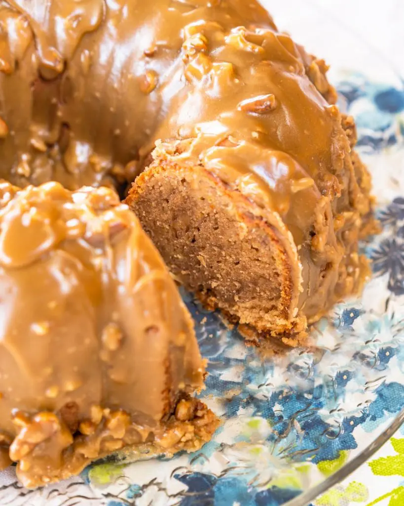Side angle of a bananas foster bundt cake with a slice missing, frosted with a caramel pecan praline glaze on a glass plate over a blue floral napkin.
