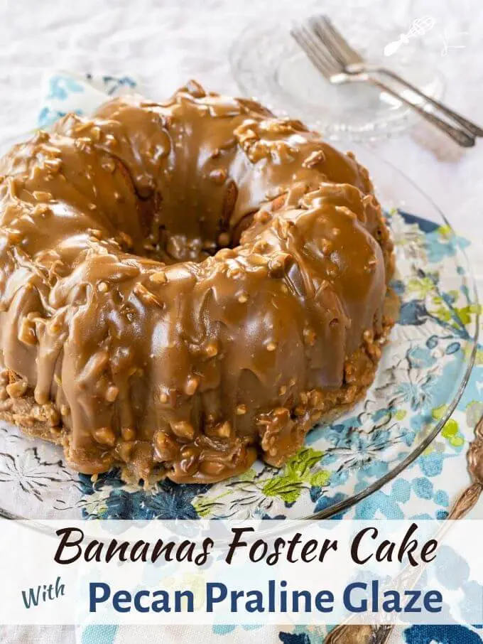 Top angle of a bananas foster bundt cake frosted with a caramel pecan praline glaze on a glass plate over a blue floral napkin. Plates, a cake server, and forks sit in the background. The title banner sits below the photo.