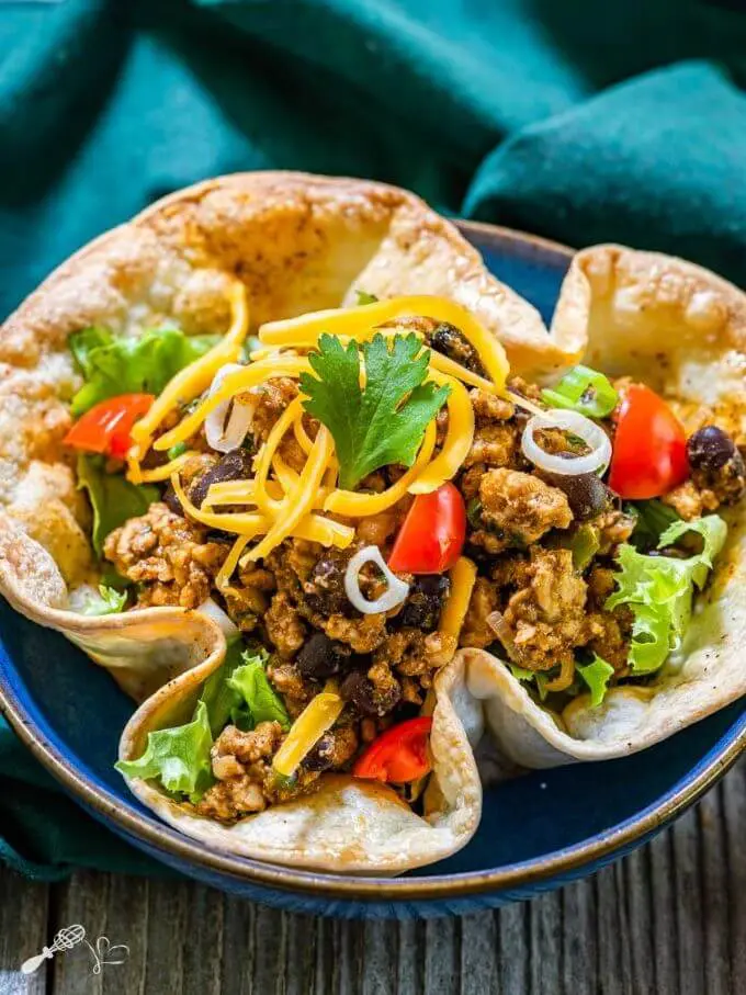 Top angel view of a taco bowl filled with spicy ground chicken and black bean filling topped with shredded cheese, lettuce and tomatoes on a green napkin.