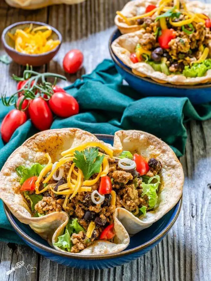 Top angel view of a taco bowl filled with spicy ground chicken and black bean filling topped with shredded cheese, lettuce and tomatoes on a green napkin. A bunch of cherry tomatoes, a bowl of shredded cheese and a partial taco salad sits in the background.