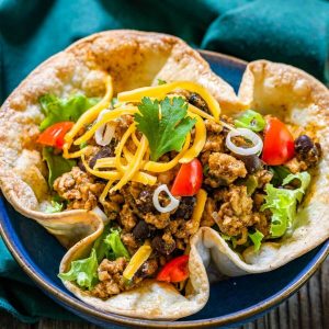 Top angel view of a taco bowl filled with spicy ground chicken and black bean filling topped with shredded cheese, lettuce and tomatoes on a green napkin.