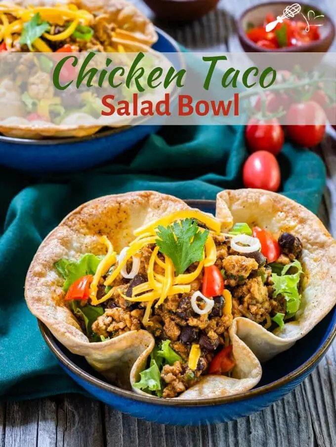 Top-down view of two taco bowls loaded with ground chicken, black beans, and lettuce garnished with tomatoes, cilantro, and shredded cheese on a green napkin. A small bowl of diced tomatoes and a bunch of fresh tomatoes sit off to the side. The title \"Chicken Taco Salad Bowl runs across the top.