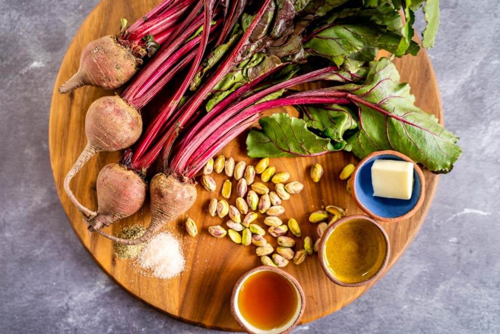 A round wooden cutting board topped with raw red beets, pistachios, dishes of honey, oil, and butter, a small pile of salt and pepper.