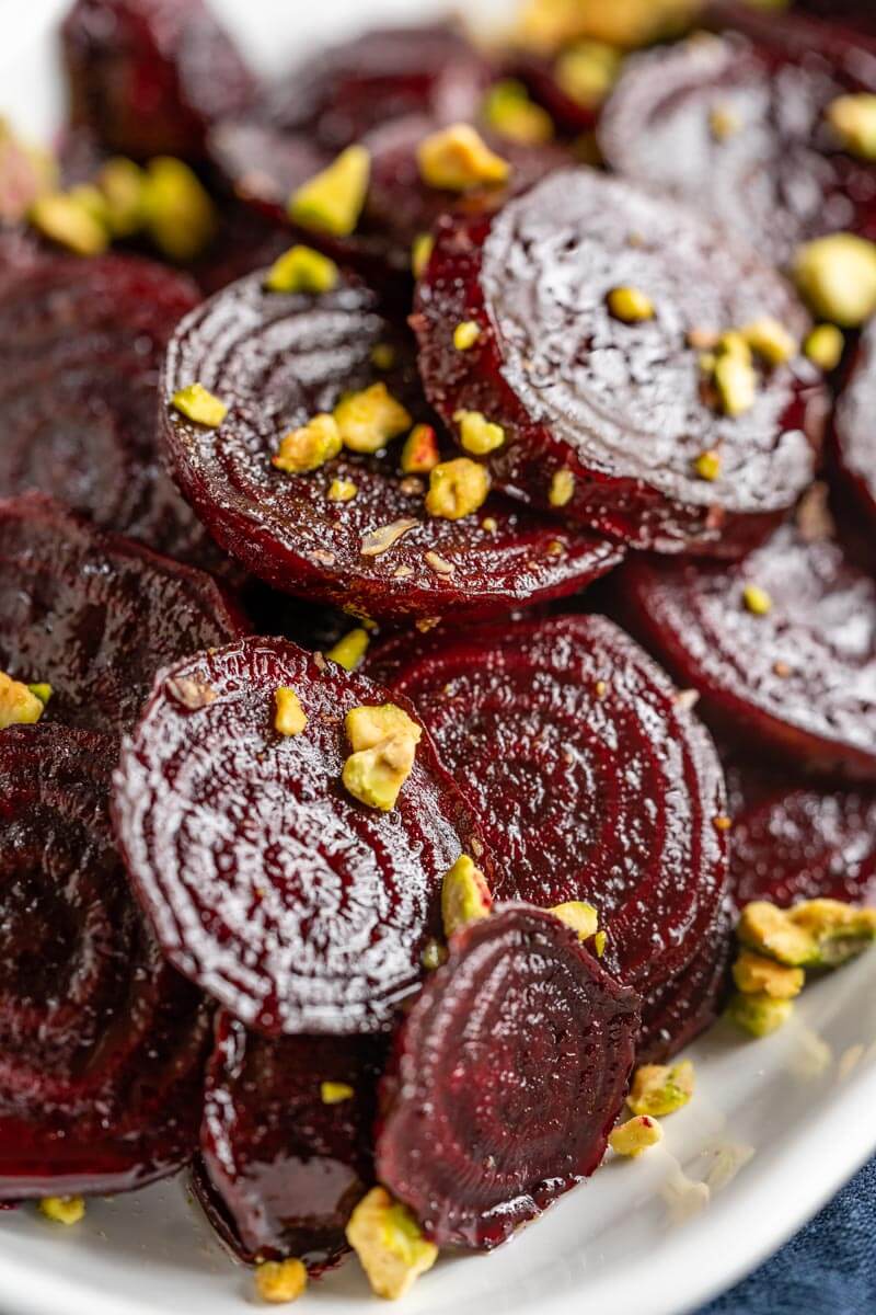 Close-up side-angle photo of glistening red beet slices in a white bowl sprinkled with chopped pistachio nuts.