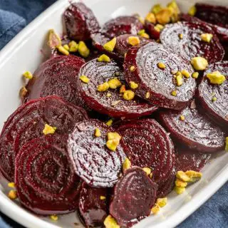 Close-up angle photo of glistening red beet slices in a white bowl sprinkled with chopped pistachio nuts. The dish is sitting on a blue napkin.