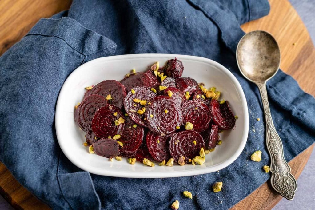 Top down photo of glistening red beet slices in a white bowl sprinkled with chopped pistachio nuts. The dish is sitting on a blue napkin next to an antique serving spoon.