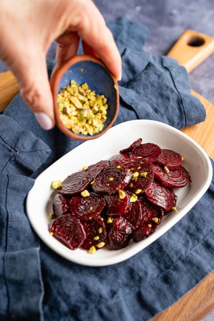 A hand holding a small dish of chopped pistachio nuts being sprinkled onto a white dish filled with glistening oven roasted sliced red beets.