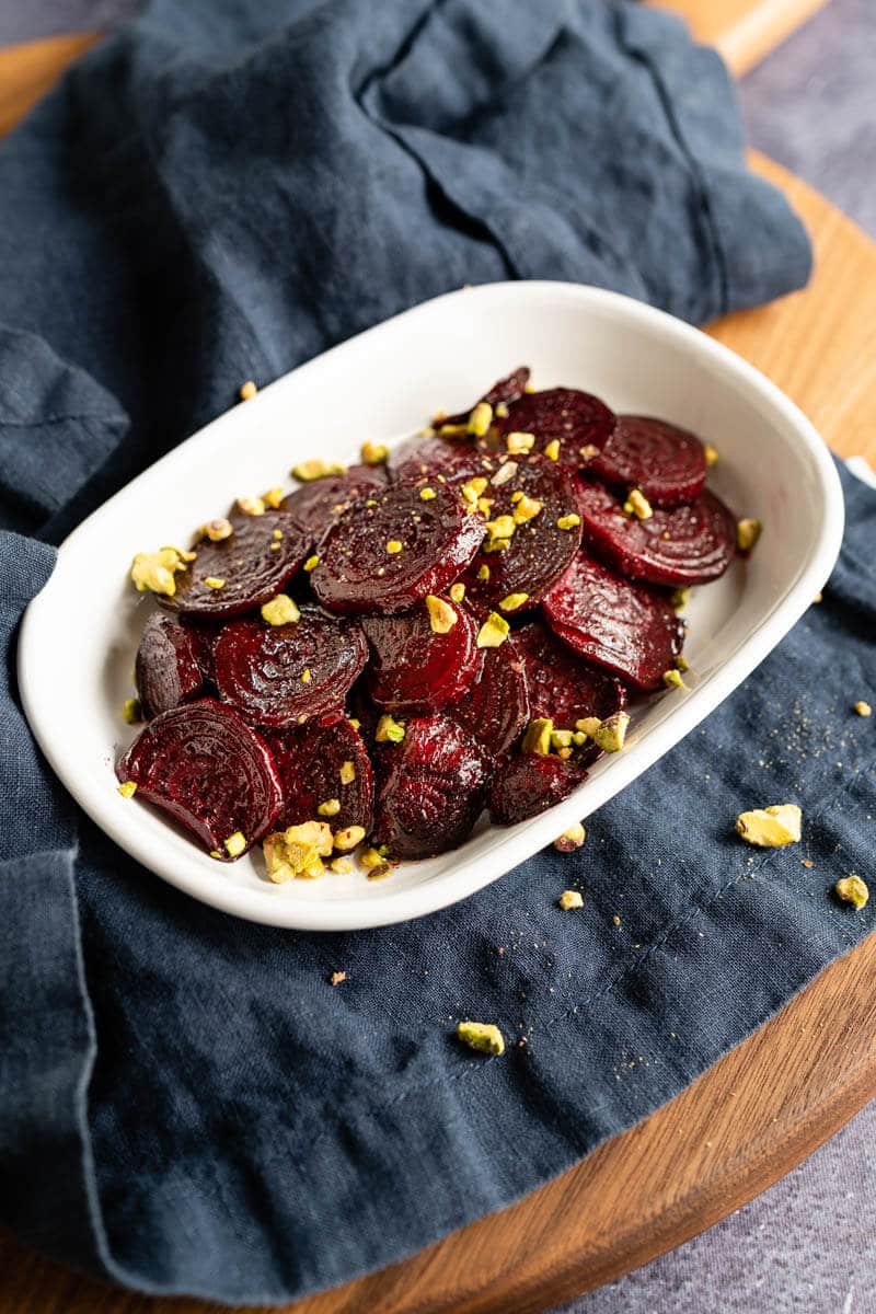 Close-up angle photo of glistening red beet slices in a white bowl sprinkled with chopped pistachio nuts. The dish is sitting on a blue napkin.