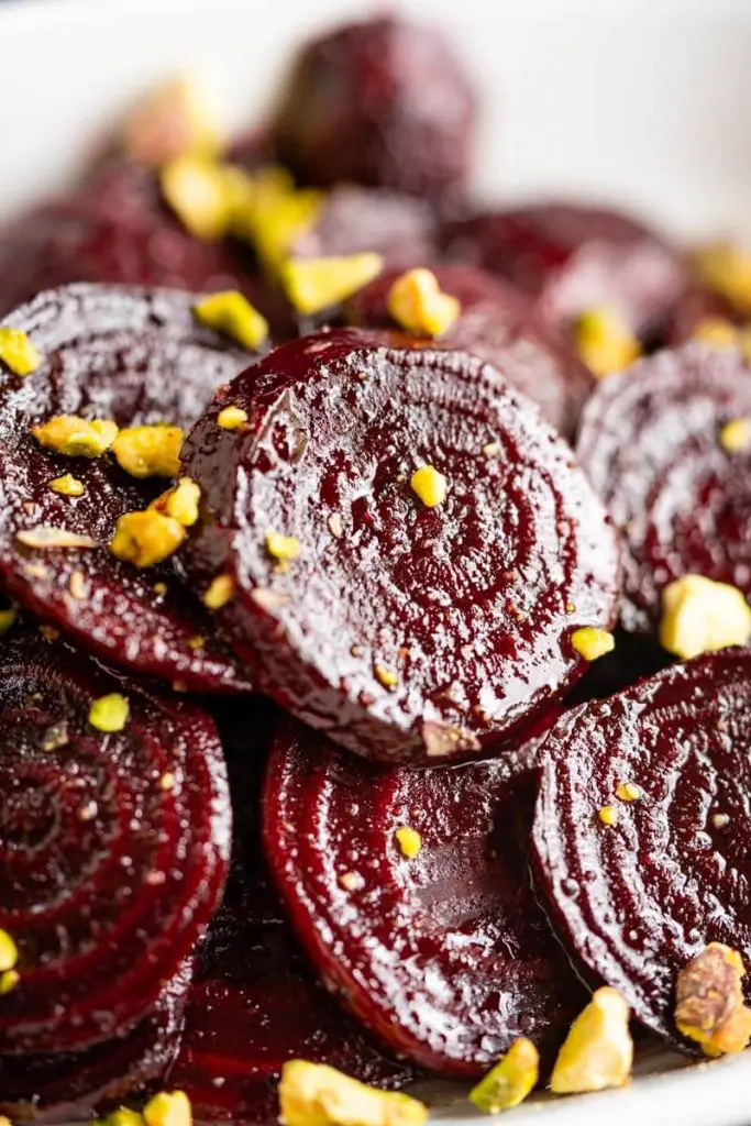 Close-up side-angle photo of glistening red beet slices in a white bowl sprinkled with chopped pistachio nuts.