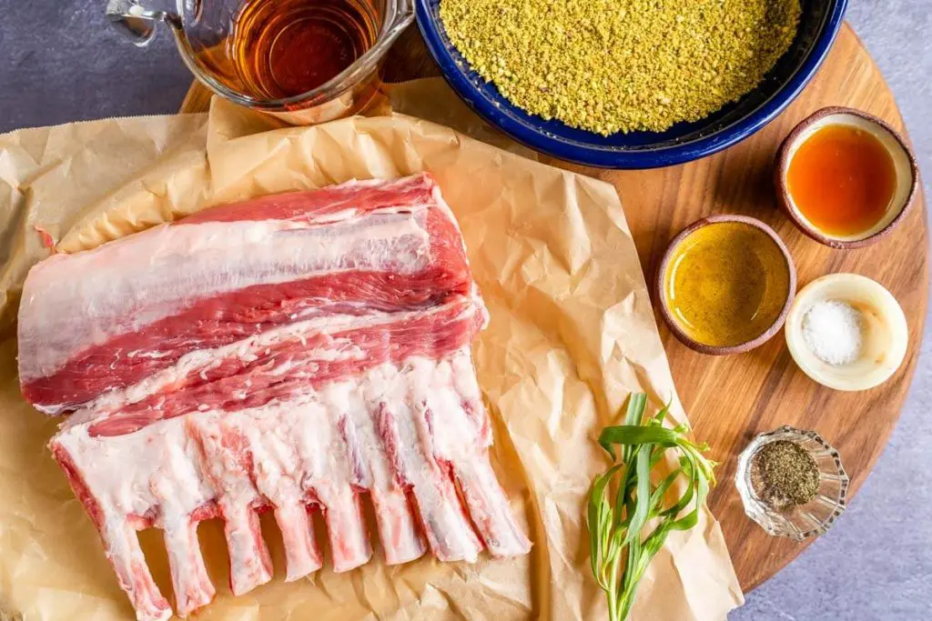 Top down photo of the ingredients used for pistachio crusted lamb chops including a rack of lamb, tarragon, salt, pepper, sherry, honey oil and chopped pistachios.