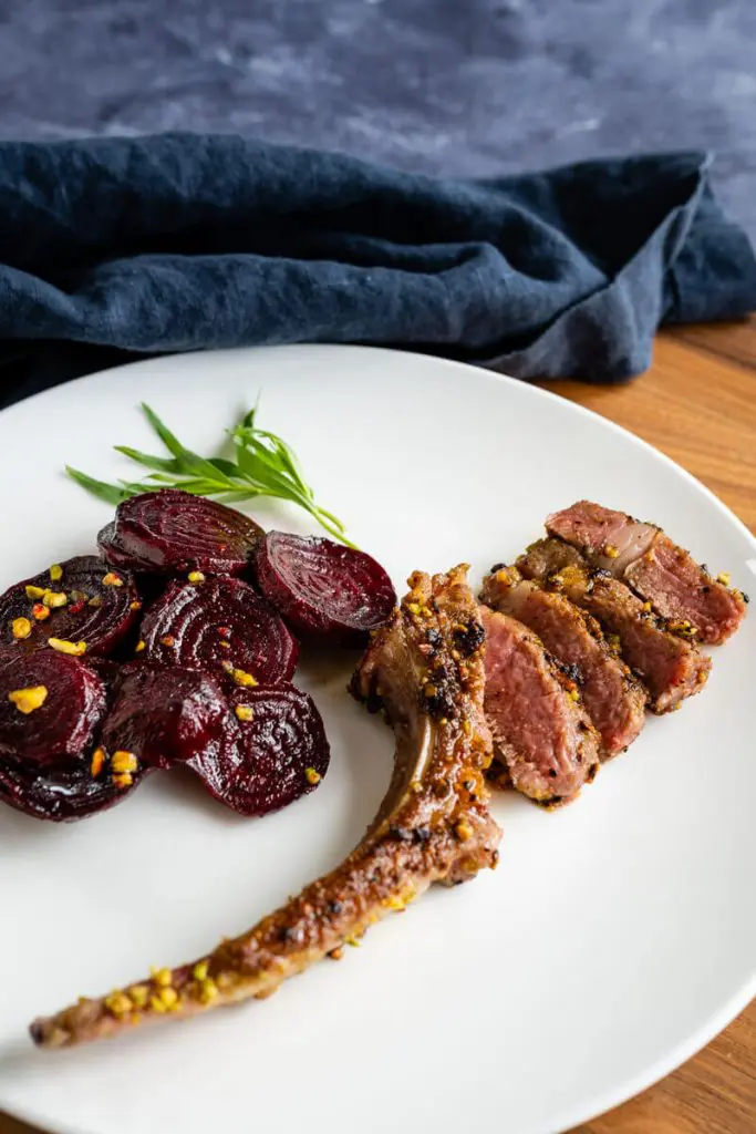 White plate showing a pan-fried pistachio crusted lamb chop next to a serving of sliced red beets sprinkled with pistachios. A sprig of tarragon is on the plate. A blue napkins sits in the background.